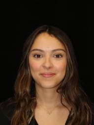 Professional headshot portrait of young woman with long straight brown hair against dark background with neutral expression, suitable for professional profiles or identification